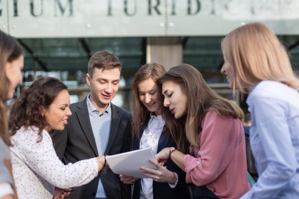 Grupa studentów przed wydziałem budynku analizuje dokument trzymany przez studenta w garniturze