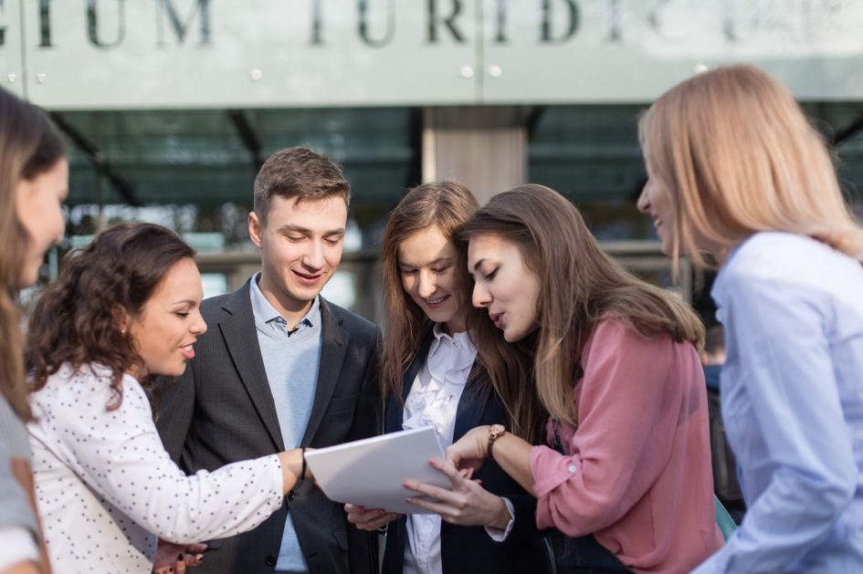 Grupa studentów przed wydziałem budynku analizuje dokument trzymany przez studenta w garniturze