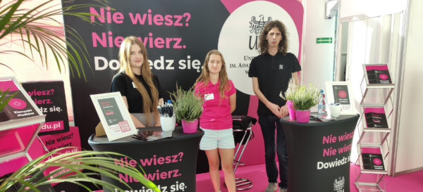 Troje studentów: dwie studentki i jeden student w uczelnianych koszulkach polo pozują do zdjęć na stoisku UAM.