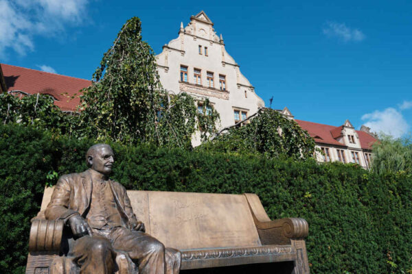 Ławeczka Heliodora Święcickiego przed Collegium Minus Uniwersytetu im. Adama Mickiewicza w Poznaniu. W tle zabytkowy gmach uczelni i zieleń kampusu pod błękitnym niebem